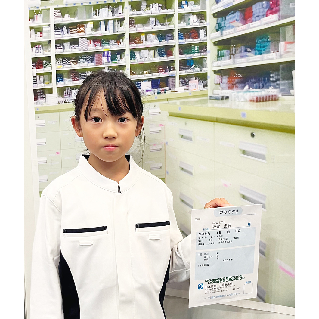 調剤室内の写真ポスターの前で日本調剤の薬剤師ユニフォームを着て薬袋を持ち、記念撮影をする女の子
